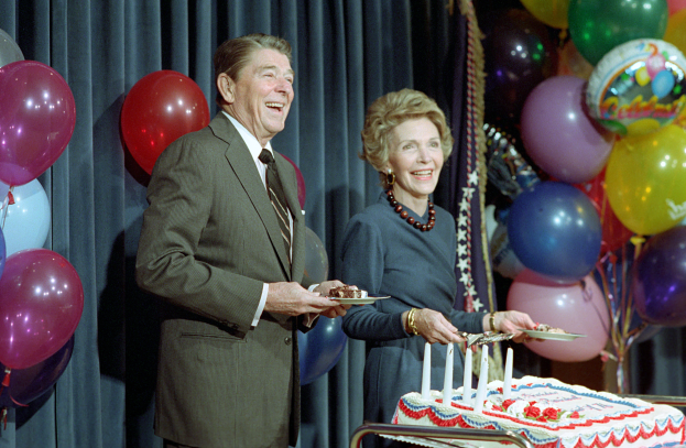 Ein Mann und eine Frau stehen nebeneinander, lächeln und halten Teller in den Händen, vor einem Tisch mit einer Kerzen geschmückten Torte, bunten Ballons und einem Vorhang im Hintergrund, feiernd ihren 60. Geburtstag.
