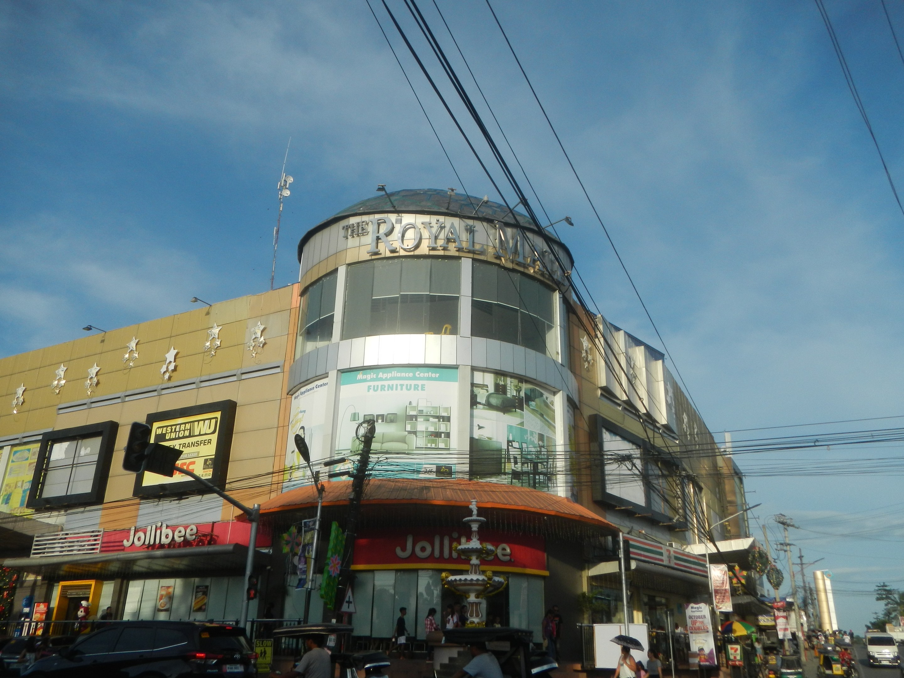 Eine belebte Stadtstraße mit Fahrzeugen auf der Straße, Fußgängern auf dem Gehweg und dem Royal Mall Einkaufszentrum im Hintergrund, umgeben von Bäumen, Strommasten und Drähten unter einem klaren Himmel.