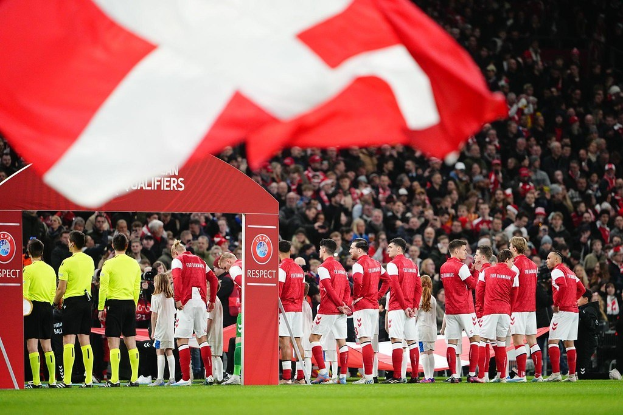 Gruppe von Menschen auf einem Fußballfeld mit einer roten und weißen Flagge im Vordergrund, einem Bogen mit "Bayern München vs Bayern München Wetten & Vorschau"-Text im Hintergrund und einer großen Menschenmenge im Stadion.