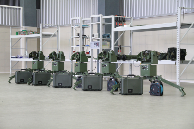 Eine Gruppe von Militärrobotern ist auf einem Boden positioniert, umgeben von Koffern und anderen Gegenständen auf Gestellen, mit einer Wand und einem Geländer im Hintergrund.