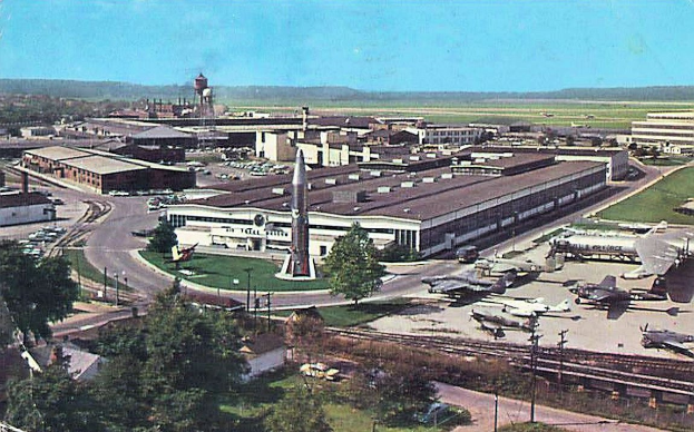 Flughafenterminal mit umliegenden Fahrzeugen, Laternen, Bäumen und einem bewölkten Himmel.