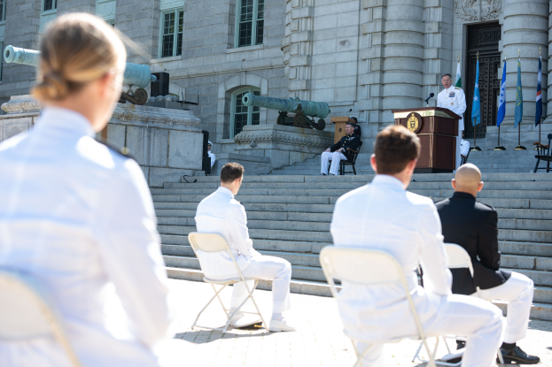Gruppe von Männern in weißen Uniformen auf Stühlen vor einem Gebäude mit Fenstern, Säulen und einer Tür sitzen, mit einer Person in der Nähe eines Podiums mit Mikrofon und einer weiteren Person im Hintergrund, zusammen mit Kanonen und Fahnenmasten, was auf eine Zeremonie an der US-Navy-Akademie hinweist.