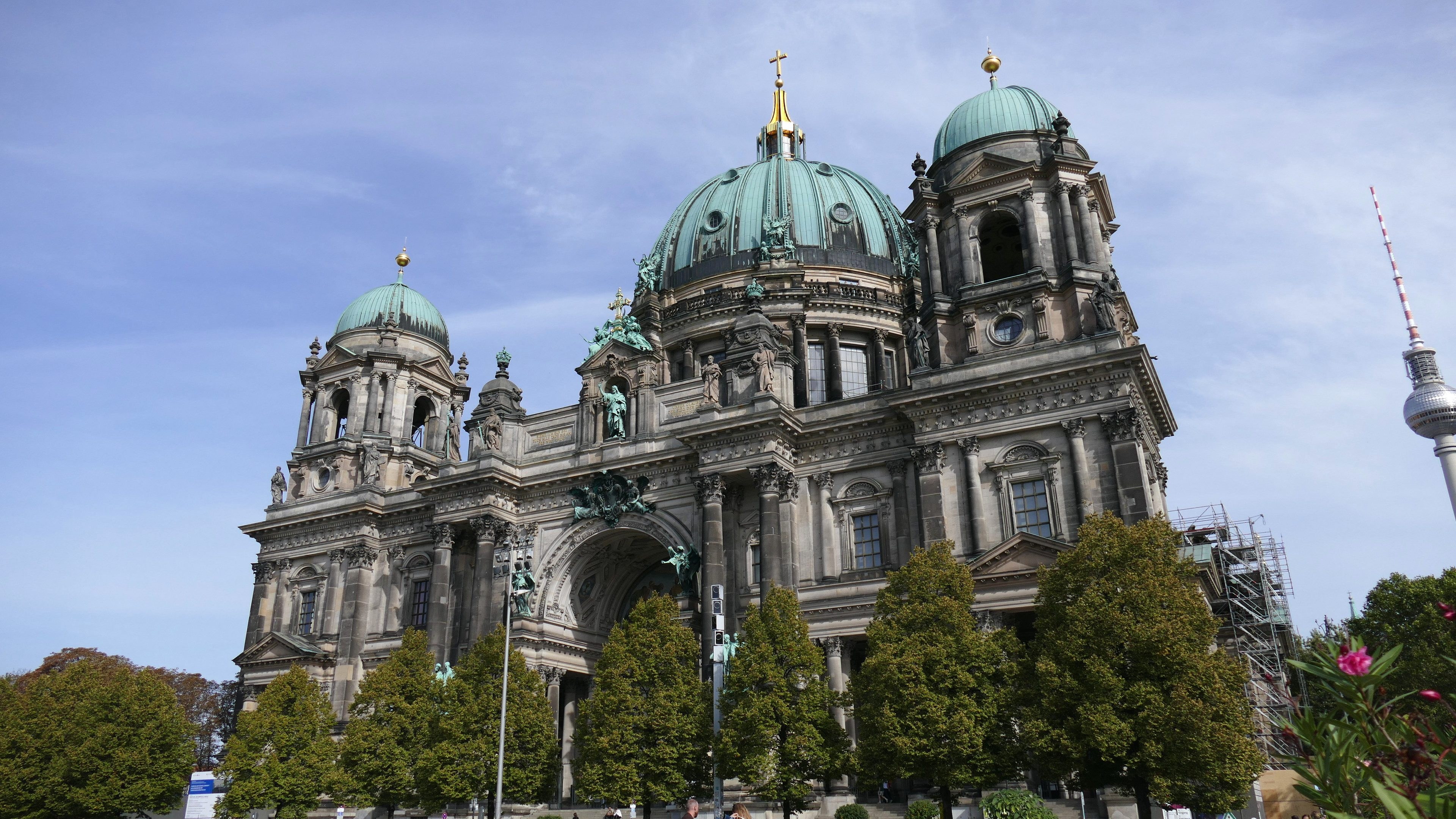 Der Berliner Dom, eine große Kathedrale mit Fenstern, Säulen, Bögen und Statuen, steht prominent in Berlin, Deutschland, mit Menschen, die davor gehen, Bäumen, Blumenpflanzen und einem Turm in der Nähe unter einem bewölkten Himmel.