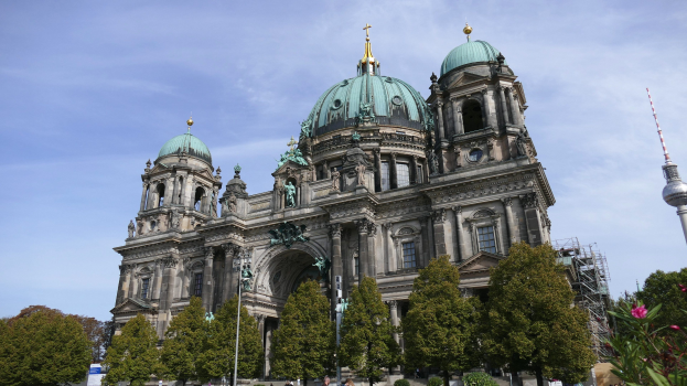 Der Berliner Dom, eine große Kathedrale mit Fenstern, Säulen, Bögen und Statuen, steht prominent in Berlin, Deutschland, mit Menschen, die davor gehen, Bäumen, Blumenpflanzen und einem Turm in der Nähe unter einem bewölkten Himmel.