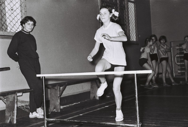 Schwarzes und weißes Foto einer Frau, die über ein Hindernis in einer Turnhalle springt, mit Zuschauern und Bänken im Hintergrund.