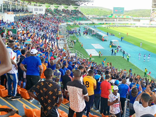 Eine große Menschenmenge versammelt sich vor einem eingezäunten Fußballfeld, wobei einige auf Stühlen sitzen und andere stehen, unter einem klaren blauen Himmel mit Hügeln im Hintergrund, in Erwartung des 2019 African Cup of Nations-Spiels.