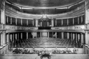 Schwarz-weiß-Foto des Theatersaals im Londoner Theatre Royal, das Sitzreihen, Treppen, Geländer, Säulen, Lampen und einen Kronleuchter mit einigen sitzenden und stehenden Menschen zeigt.