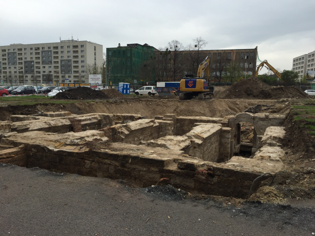 Großer Erdberg an einer Baustelle umgeben von Baggern, Fahrzeugen, Gras, Bäumen, Gebäuden und einer Straße unter einem bewölktem Himmel.