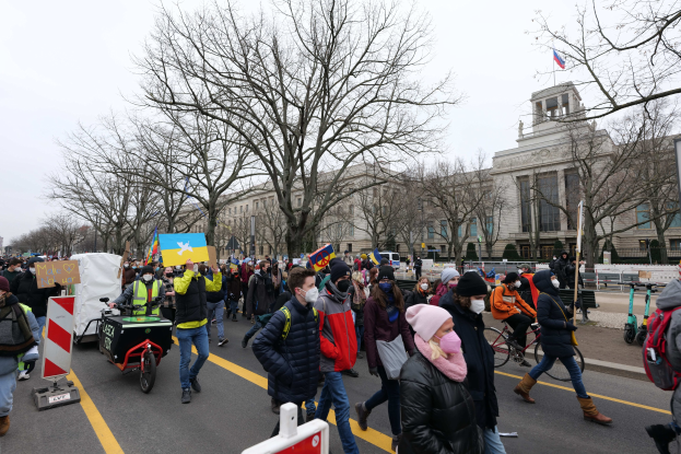 Eine große Gruppe von Menschen marschiert auf einer Stadtstraße in Washington, D.C. und hält Schilder und Banner hoch, während einige Fahrräder fahren, unter einem klaren blauen Himmel.