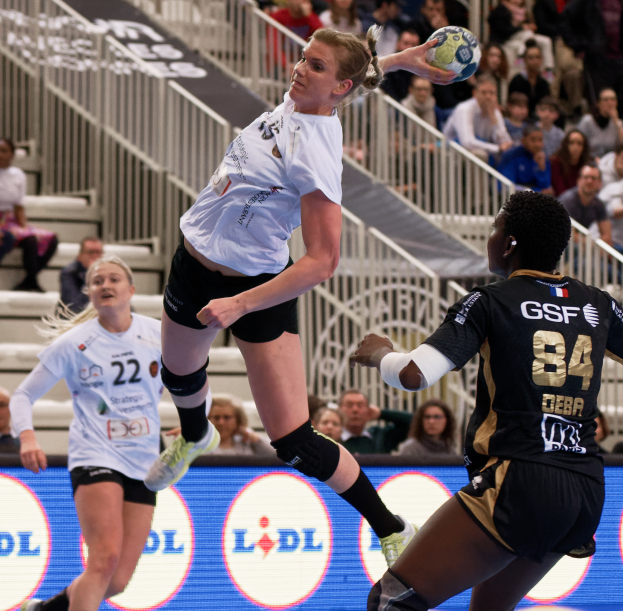 Eine Gruppe von Frauen, die auf einem Platz Handball spielen, mit einem Ball in der Luft, Zuschauern auf Treppen und Geländern im Hintergrund und einem Schild mit der Aufschrift "Handball-Weltmeisterschaft 2019 - Tag 2".