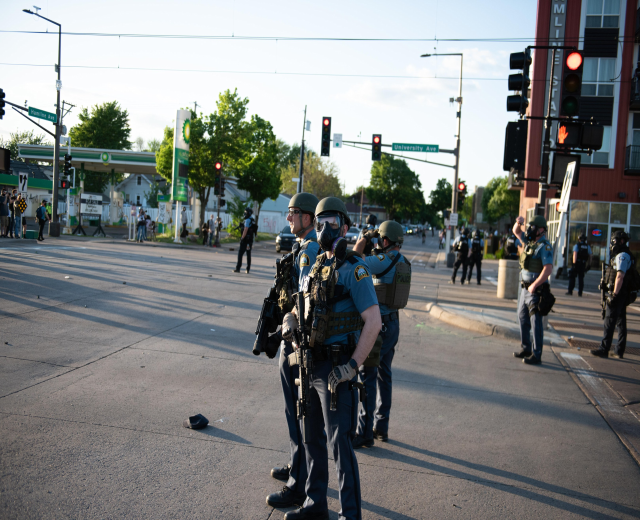 Gruppe von Polizisten in Helmen und Ausrüstung auf einer Straße mit Fahrzeugen, Verkehrszeichen, Schildern, Bäumen, Gebäuden und einem bewölkten Himmel im Hintergrund.
