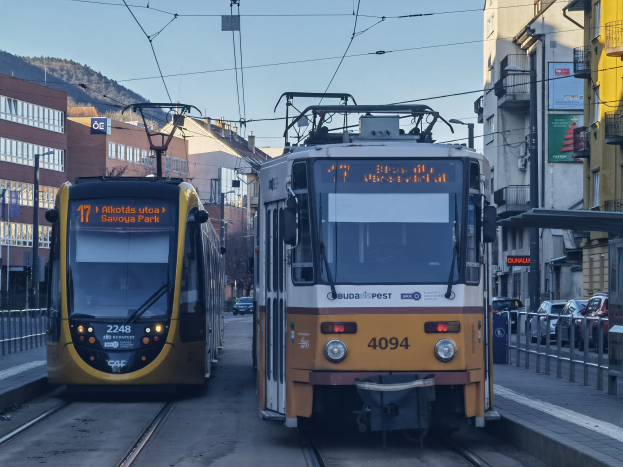 Zwei gelbe und weiße Straßenbahnen fahren auf Stadtbahngleisen neben hohen Gebäuden, mit Fahrzeugen auf der Straße, Geländern auf beiden Seiten und einem Hintergrund mit Gebäuden, Polen, Schildern, Bäumen, einem Hügel und dem Himmel.