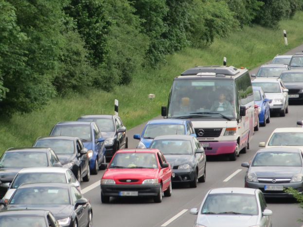 Verkehrsstau auf einer Autobahn mit mehreren Autos und einem Lieferwagen, Menschen sichtbar in Fahrzeugen, Bäume und Gras im Hintergrund.