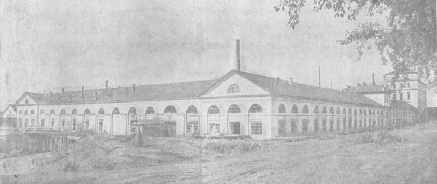 Schwarz-weiß-Fotografie eines großen, mehrstöckigen Gebäudes mit zahlreichen Fenstern, umgeben von Grün und Bäumen, vor einem klaren Himmel, mit der Aufschrift "1892 – das erste Fabrikgebäude in der Stadt" unten.