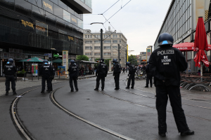 Eine Gruppe von Polizeibeamten in schwarzen Uniformen und Helmen steht auf einer Straße, die von Glasfenster-Gebäuden gesäumt ist, mit Bäumen, Strommasten und verstreuter Außenmöblierung im Hintergrund unter einem klaren Himmel.
