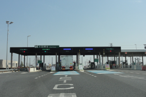 Mautstation mit einem vor ihr geparkten Lastwagen auf einer Straße mit weißen und blauen Markierungen, Laternen, Schildern und Gebäuden im Hintergrund unter einem klaren blauen Himmel.