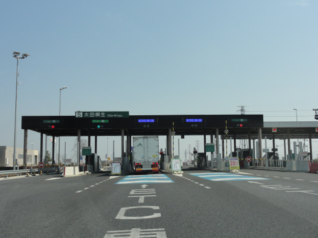 Mautstation mit einem vor ihr geparkten Lastwagen auf einer Straße mit weißen und blauen Markierungen, Laternen, Schildern und Gebäuden im Hintergrund unter einem klaren blauen Himmel.