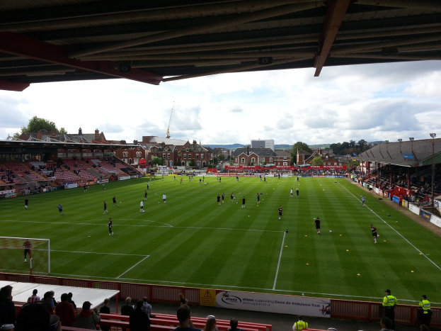 Ein Fußballspiel im St. Mary's Stadium, mit Zuschauern auf den Tribünen und in der Nähe, einem Torpfosten auf der linken Seite und im Hintergrund Gebäude, Bäume und ein bewölkter Himmel.