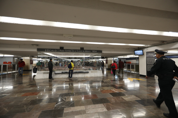 Eine Gruppe von Menschen geht durch eine belebte U-Bahn-Station mit einem Polizeibeamten im Vordergrund, überirdische Schilder und Lichter sowie verschiedene Gegenstände wie Geländer und Taschen.