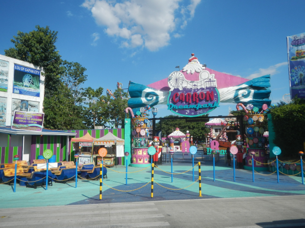 Eine Karussellfahrt am Parkeingang mit einer Straße, einem Zaun, Buden, Schildern, Bäumen und einem bewölkten Himmel im Hintergrund.