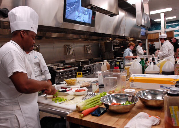 Eine Gruppe von Köchen in einer Küche beim Zubereiten von Essen auf einem Schneidbrett, wobei ein Koch im Vordergrund in einer weißen Schürze und Mütze mit einem Messer in der Hand zu sehen ist und von Utensilien, Schüsseln, Gemüse, einem Mobiltelefon, Flaschen und anderen Gegenständen umgeben ist, während im Hintergrund ein Fernseher und Deckenlampen zu sehen sind.