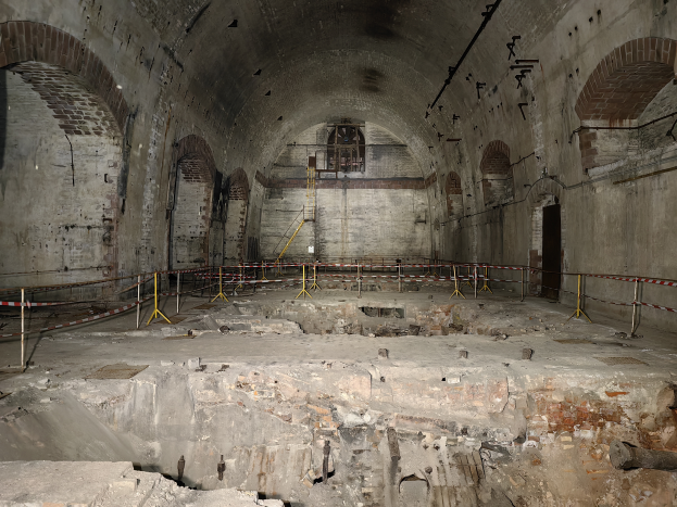 Ein altes Gebäude mit Schutt auf dem Boden, umgeben von Gittern und Pfählen, mit einer Wand mit Bögen und einem Fenster im Hintergrund.