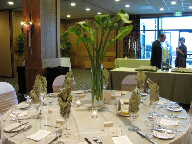 Ein Hotelesszimmer mit einem weißgedeckten Tisch, der mit Tellern, Besteck und Gläsern gedeckt ist, umgeben von Stühlen, mit einem grünen Tisch im Hintergrund, einem Mann in einem schwarzen Blazer, einem Mädchen neben ihm und einer Tür mit der Aufschrift "Ausgang" und zwei Männern dahinter.