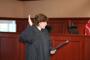 Eine Frau in einer schwarzen Robe steht an einem Richtertisch und hält eine Akte in der Hand, mit einem Mikrofon und einer Flagge auf dem Tisch, Stühle dahinter und eine fensterverglaste Wand im Hintergrund.