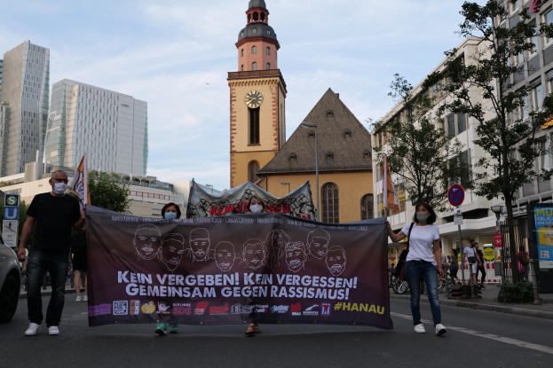 Eine Gruppe von Menschen in Masken, die eine Straße entlanggehen und ein Banner mit Text und Bildern halten, mit einem geparkten Auto links daneben, Gebäuden, Bäumen, Schildern, Pfosten, einem Uhrenturm und einem klaren blauen Himmel im Hintergrund.