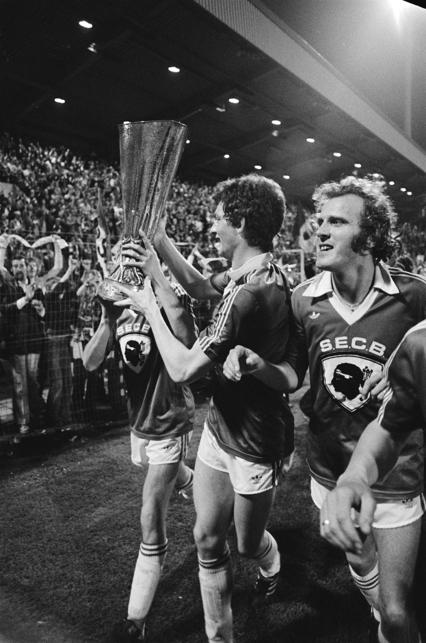 Schwarzes Foto von Fußballspielern, die mit einem Pokal auf einem Feld feiern, umgeben von einer Menge hinter einem Zaun unter Stadionbeleuchtung.