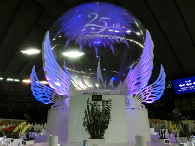 Ein großer Kristallball mit Flügeln oben drauf, umgeben von Blumenschmuck, Namensschildern und anderen Gegenständen auf dem Boden, vor einer dekorierten Kulisse mit Schildern, Lichtern und einem Bildschirm für eine 25-jährige Jubiläumsfeier.