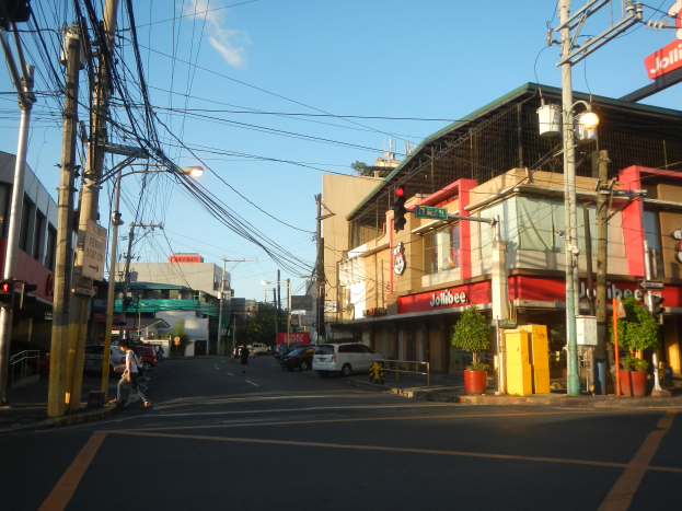 Stadtstraße mit Autos, Fußgängern, Strommasten, Verkehrszeichen, Gebäuden mit Schildern, Topfpflanzen und bewölktem Himmel.