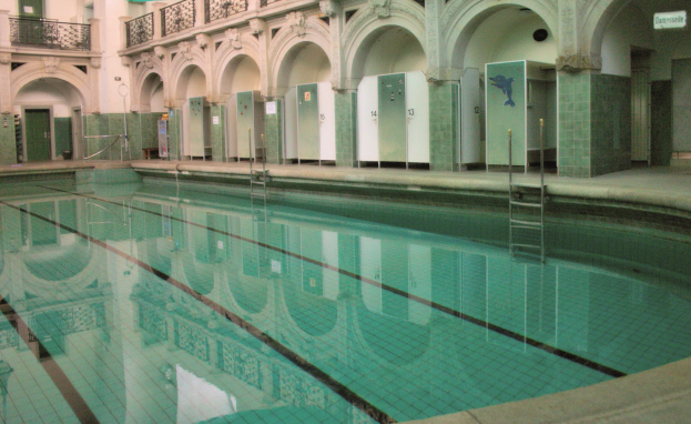 Innenbad mit klarem Wasser, umgeben von gefliestem Boden, Bogengängen, Säulen, Geländern, Türen und Texttafeln in einem großen Gebäude.