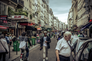 Zwei große Gebäude mit einer Straße dazwischen, wo eine Menge Menschen geht und mehrere Stände auf dem Erdgeschoss eingerichtet sind, mit einigen Mülltonnen sichtbar. Ein Mann fährt ein Fahrrad auf der Straße, und Autos sind auf der gegenüberliegenden Seite geparkt.