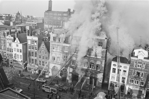 Schwarzes und weißes Foto einer Stadtstraße mit einem Feuer in der Mitte, umgeben von Gebäuden, Fahrzeugen und Menschen, mit Rauch in der Luft und dem Himmel im Hintergrund.