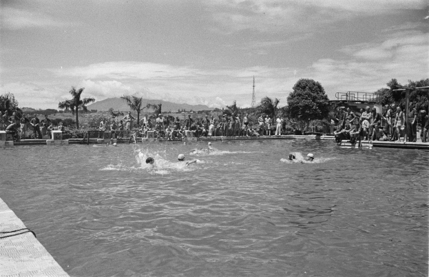 Schwarz-weiß-Foto von Schwimmern in einem Pool mit Zuschauern, einem Zaun, Pfählen, einem Turm, Bäumen, Hügeln und einer bewölkten Himmel.