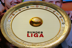 Ein Mann hält ein goldenes Schild mit 'Bundesliga' hoch, im Hintergrund eine Fahne mit drei Personen.