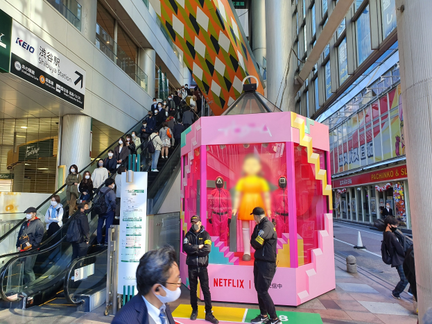 Eine Gruppe von Menschen steht vor einem Gebäude mit Glaswänden, Säulen und Texttafeln, einige tragen Masken, während eine Rolltreppe mit Menschen zu sehen ist. In der Mitte befindet sich ein pinker Kasten, der mit 'Netflix's new Netflix booth in Tokyo' beschriftet ist und Puppen enthält.