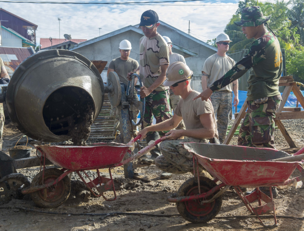 Gruppe von Männern mit Helmen auf einer Baustelle, einige mit Handwagen und einer Person mit Betonmischer, mit Gebäuden, Bäumen, Strommasten und einem bewölkten Himmel im Hintergrund.