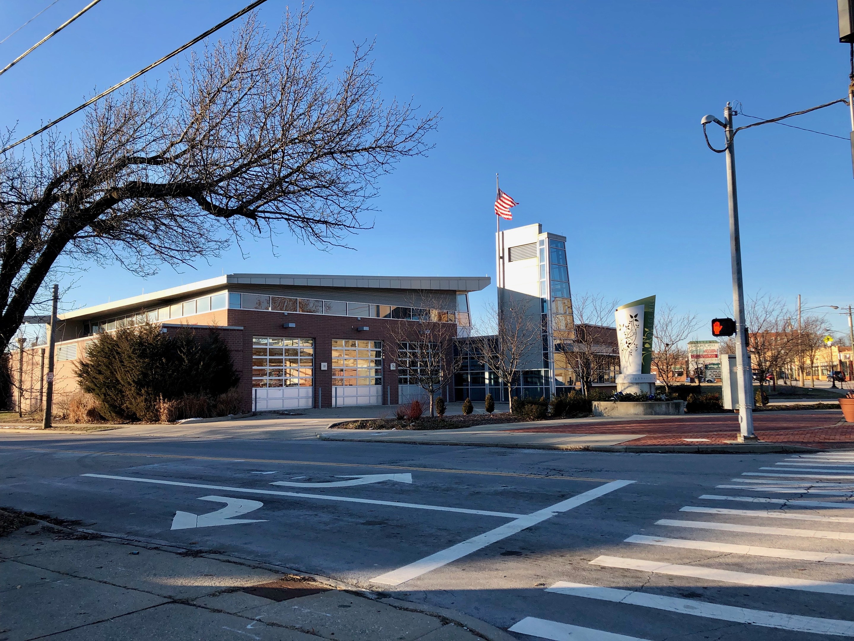 Großes Gebäude mit einer Flagge oben drauf, als neue Feuerwache an einer Straßenkreuzung identifiziert, umgeben von Bäumen, Straßenmöbeln und sichtbarem Himmel.