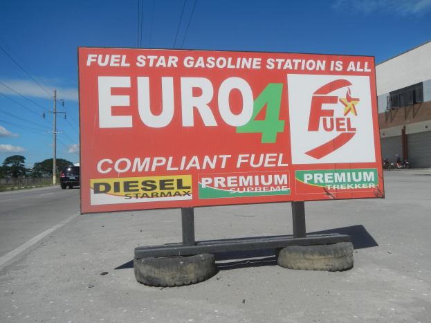 Tankstelle mit "Euro4 Fuel"-Schild, Fahrzeuge auf der Straße, Strommasten, Bäume, ein Gebäude auf der rechten Seite und sichtbarer Himmel.
