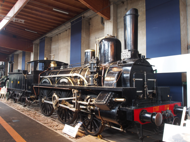 Alte Dampflokomotive im Museum ausgestellt, umgeben von Eisenbahnschienen und Informationsschildern, mit einer Wand und beleuchteter Decke im Hintergrund.