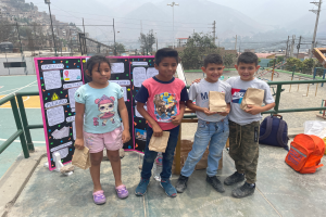 Eine Gruppe von Kindern steht auf einem Basketballfeld, jedes hält einen Papiertüte, mit Tüten und Gegenständen rechts und einer Tafel mit Text links, mit Zaun, Pfosten, Straßenlaternen, Strommasten mit Drähten, Bäumen, Gebäuden, Hügeln und Himmel im Hintergrund.