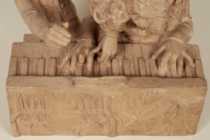 Eine steinerne Skulptur eines gekrönten Mannes in einem langen Gewand, der konzentriert ein Piano spielt, vor einem weißen Hintergrund.