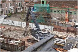 Ein Bagger arbeitet auf einer Baustelle umgeben von Gebäuden, Bäumen, Pflanzen, Pfählen, einem Zaun und Fahrzeugen auf einer Straße, mit einem Container mit Gegenständen in der Nähe und Text am unteren Bildrand.