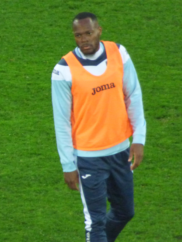 Ein Fußballer in einem orangen Trikot und blauen Hosen läuft entschlossen über ein grünes Feld.