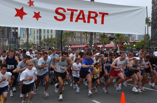 Gruppe von Läufern bei einem Marathon, die an einer Verkehrskegel vorbei laufen, mit einem Banner und Gebäuden im Hintergrund unter einem klaren blauen Himmel.