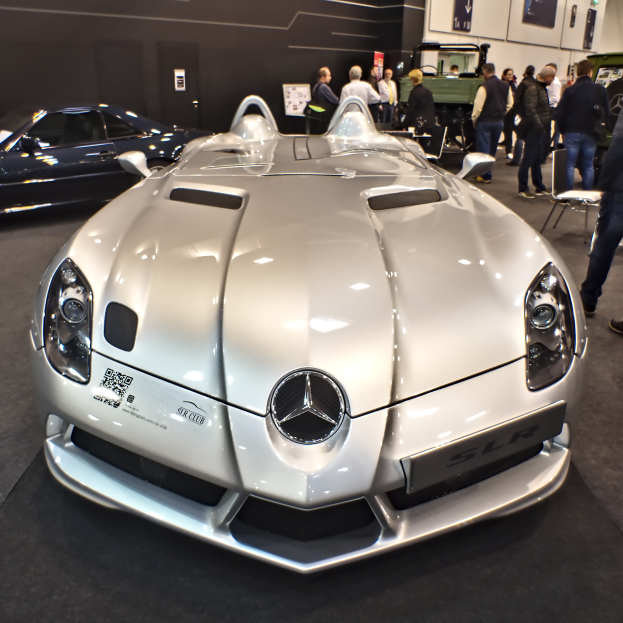 Ein Mercedes-Benz SLR McLaren Roadster wird auf dem Genfer Automobilsalon präsentiert, umgeben von mehreren Bewunderern, während die Ausstellungswände mit Plakaten und Lichtern geschmückt sind.