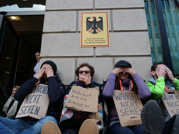 Eine Gruppe von Menschen sitzt vor einem Gebäude und hält Schilder, wobei einige ihre Gesichter verdecken, scheinbar protestierend.