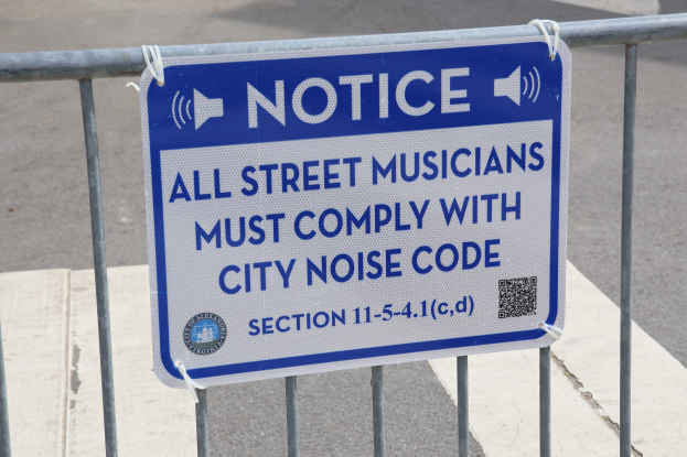 Hinweistafel an einem Zaun mit einem Schild, auf dem "Hinweis: Alle Straßenmusiker müssen die Stadtlärmvorschriften einhalten" steht und eine Straße im Hintergrund.
